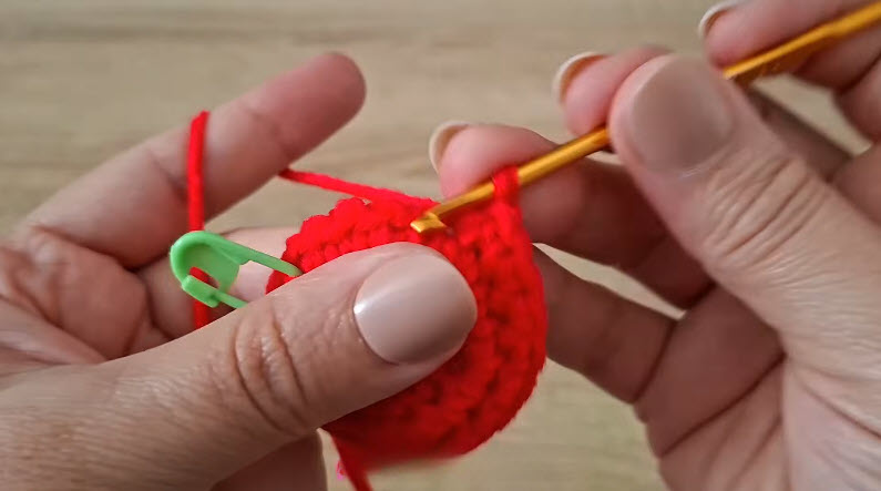 Red amigurumi piece with green stitch marker, hook mid-stitch.